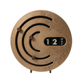 A round, disc-shaped desk clock with a brushed bronze finish, featuring an engraved leaf pattern, winding track design, and a black date window showing 12 MAY. It is supported by two small metal legs on a clean white background.